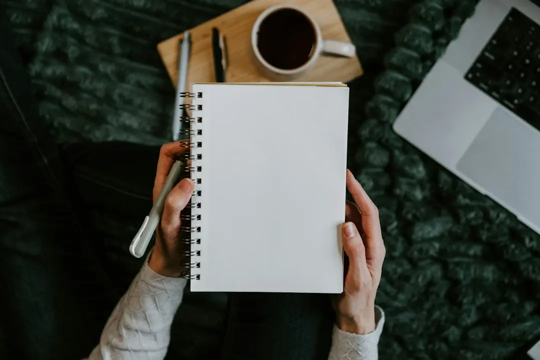 Hands holding a blank spiral notebook beside a coffee cup and pens, ready for a checklist to compare IPTV services like Nartiel8k