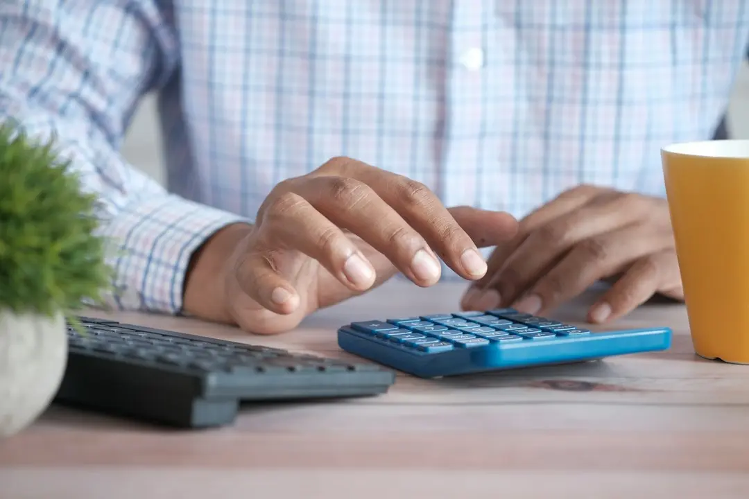 Hands typing on a blue calculator beside a keyboard and a yellow mug, working out what Nartiel8k IPTV costs per month
